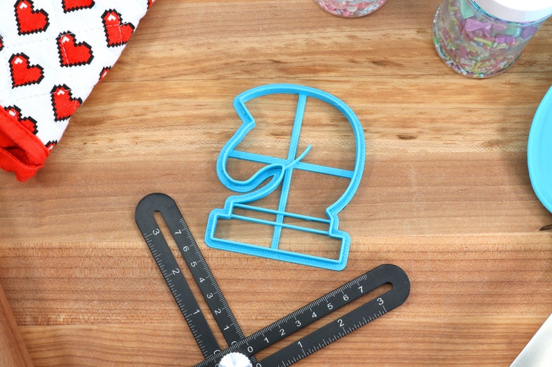 May include: A blue plastic cookie cutter shaped like a chess knight. The cutter is on a wooden surface with a ruler and a container of sprinkles.