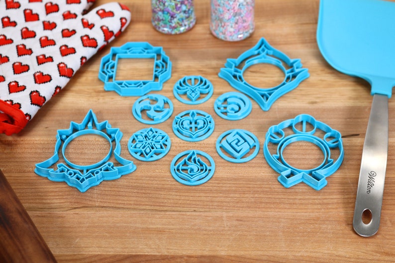 May include: A set of blue plastic cookie cutters featuring various geometric and abstract designs. The cutters are arranged on a wooden surface.