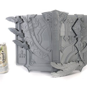 May include: A gray, intricately detailed, 3D printed book with a feather bookmark. The book is closed and has a decorative clasp. The book is about the size of a standard can of soda.