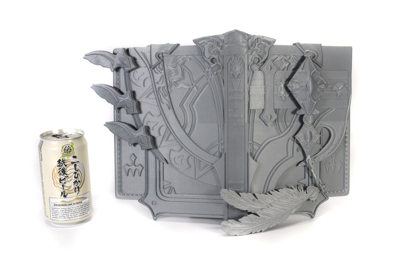 May include: A gray, intricately detailed, 3D printed book with a feather bookmark. The book is closed and has a decorative clasp. The book is about the size of a standard can of soda.