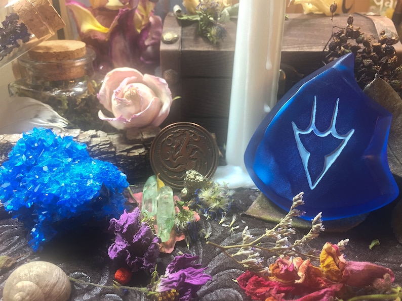 May include: A blue glass talisman with a white symbol, a copper coin with a design, a blue crystal, and dried flowers on a wooden surface.