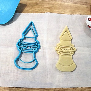 May include: A blue plastic cookie cutter and a light brown sugar cookie in the shape of a tower with a pointed top and a chain design around the middle.