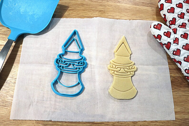 May include: A blue plastic cookie cutter and a light brown sugar cookie in the shape of a tower with a pointed top and a chain design around the middle.