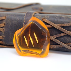 May include: An amber-colored, irregularly shaped pendant with three vertical, lighter-colored lines. The pendant is set against a backdrop of a dark brown leather journal with stitched detailing. The overall aesthetic is rustic and natural.