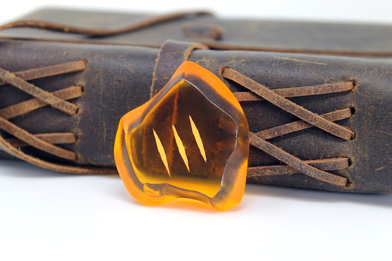 May include: An amber-colored, irregularly shaped pendant with three vertical, lighter-colored lines. The pendant is set against a backdrop of a dark brown leather journal with stitched detailing. The overall aesthetic is rustic and natural.