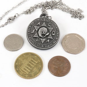 May include: A silver-colored pendant necklace with a circular pendant featuring an engraved design and text. The pendant is on a silver chain. Several coins of different sizes and colors are arranged around the pendant.