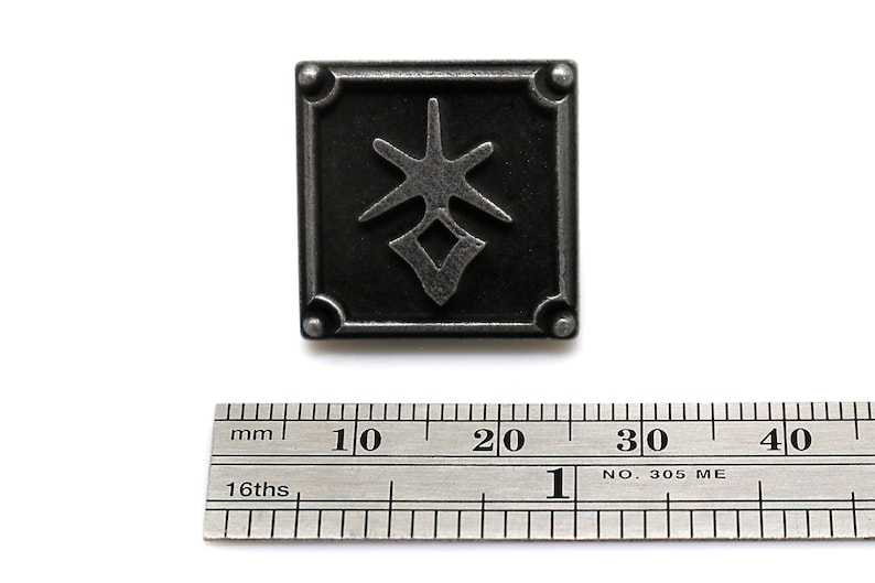 May include: Square, dark gray metal button with a stylized star and diamond design. The button is approximately 1 inch square, with rounded corners. A ruler is included for scale.