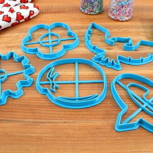May include: A collection of six bright blue plastic cookie cutters in various shapes, including a flower, leaf, gear, and abstract designs. The cutters are arranged on a wooden surface, with a red and white heart-patterned oven mitt in the background.