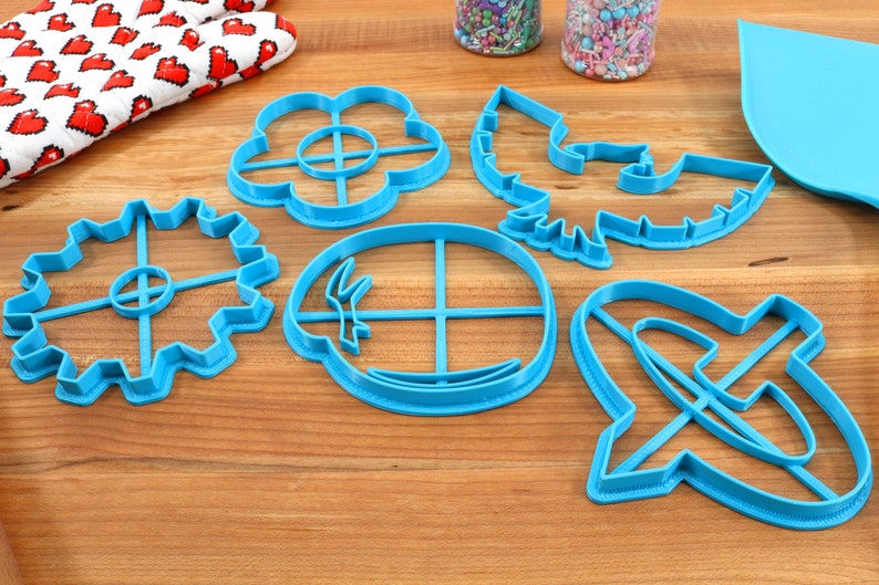 May include: A collection of six bright blue plastic cookie cutters in various shapes, including a flower, leaf, gear, and abstract designs. The cutters are arranged on a wooden surface, with a red and white heart-patterned oven mitt in the background.