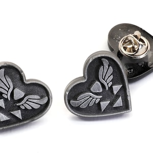 Puede incluir: Tres pines de coraz&oacute;n plateados con un s&iacute;mbolo alado y un dise&ntilde;o triangular. Los pines tienen un fondo negro y un acabado plateado.