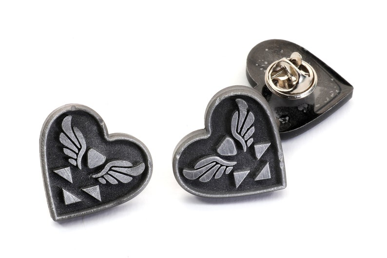 Puede incluir: Tres pines de coraz&oacute;n plateados con un s&iacute;mbolo alado y un dise&ntilde;o triangular. Los pines tienen un fondo negro y un acabado plateado.
