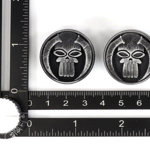 May include: Two silver-colored metal skull-shaped buttons with a black background. The skull has a detailed design with a prominent jawline and two eyes.