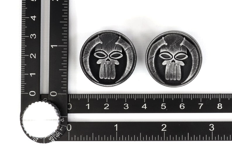 May include: Two silver-colored metal skull-shaped buttons with a black background. The skull has a detailed design with a prominent jawline and two eyes.