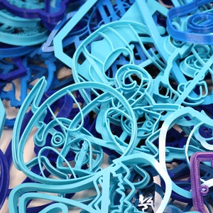 Op de afbeelding: Een close-up van een stapel blauwe en paarse plastic koekjesvormen. De vormen zijn in verschillende vormen, waaronder sterren, harten en dieren.
