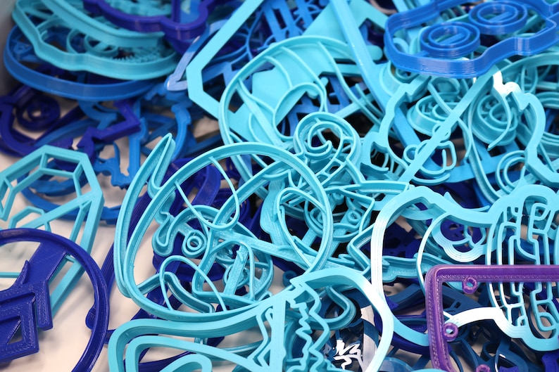 Op de afbeelding: Een close-up van een stapel blauwe en paarse plastic koekjesvormen. De vormen zijn in verschillende vormen, waaronder sterren, harten en dieren.