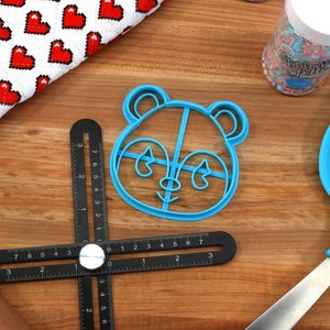 May include: A blue plastic panda cookie cutter on a wooden surface with a black ruler, a rolling pin, a spatula, and a jar of sprinkles.