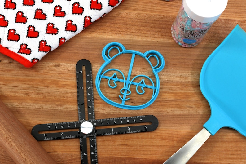 May include: A blue plastic panda cookie cutter on a wooden surface with a black ruler, a rolling pin, a spatula, and a jar of sprinkles.