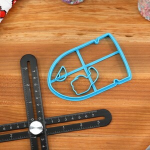 May include: A blue plastic cookie cutter shaped like a teacup with a handle. The cutter is on a wooden surface with a black ruler.