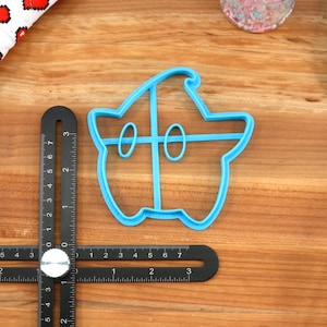 May include: A blue plastic cookie cutter shaped like a ghost with a hat. The cutter is on a wooden surface with a ruler and a spatula.