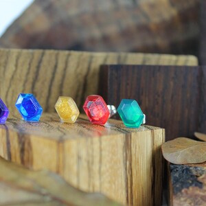 May include: Five small, faceted, gemstone-like objects in various colors, including purple, blue, gold, red, and green, are arranged on a wooden surface.