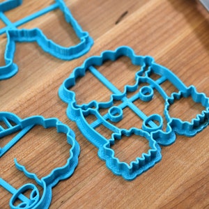 May include: Three blue plastic cookie cutters shaped like a snowman, a reindeer, and a Christmas tree. The cutters are on a wooden surface.