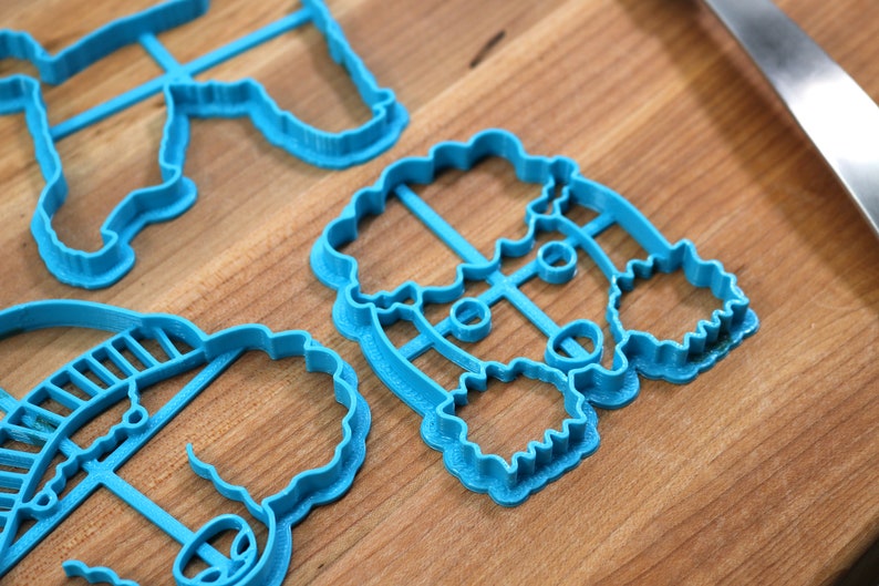 May include: Three blue plastic cookie cutters shaped like a snowman, a reindeer, and a Christmas tree. The cutters are on a wooden surface.
