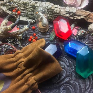 May include: A brown leather pouch with a drawstring closure, filled with four colorful gemstone-shaped objects. The pouch is surrounded by a variety of natural elements, including a bird's foot, berries, and flowers.
