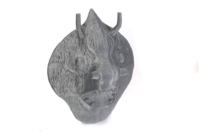 Puede incluir: Una m&aacute;scara gris impresa en 3D con una expresi&oacute;n feroz. La m&aacute;scara tiene una superficie texturizada y presenta una boca grande abierta con dientes afilados, dos ojos grandes y dos cuernos en la parte superior.