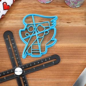 Peut inclure: Un emporte-pi&egrave;ce en plastique bleu en forme de personnage de dessin anim&eacute; avec des lunettes et une blouse de laboratoire. Une r&egrave;gle d'angle r&eacute;glable noire, une spatule bleue et un gant de cuisine rouge et blanc &agrave; motifs de c&oelig;urs sont &eacute;galement visibles.