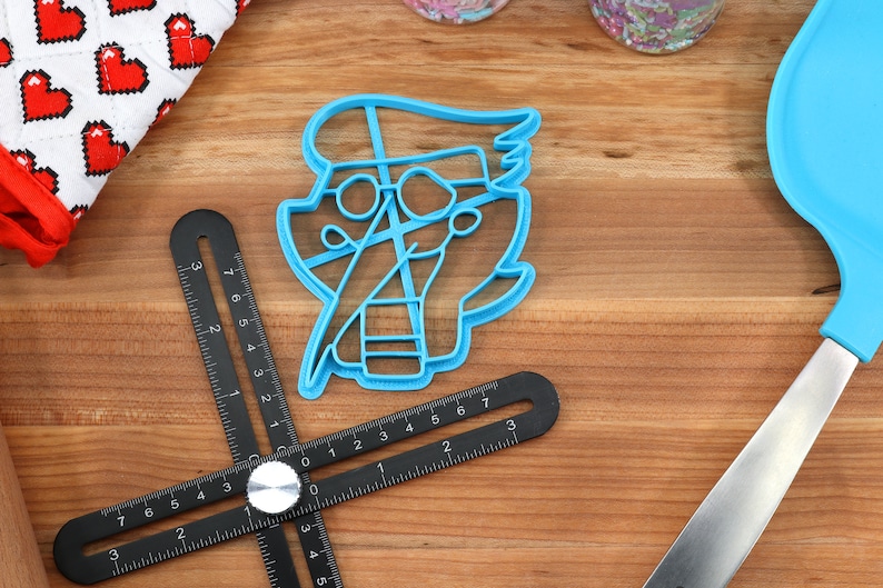 Peut inclure: Un emporte-pi&egrave;ce en plastique bleu en forme de personnage de dessin anim&eacute; avec des lunettes et une blouse de laboratoire. Une r&egrave;gle d'angle r&eacute;glable noire, une spatule bleue et un gant de cuisine rouge et blanc &agrave; motifs de c&oelig;urs sont &eacute;galement visibles.