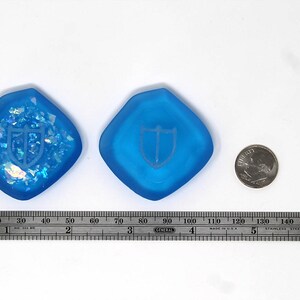 Puede incluir: Dos dados de resina de color azul, con forma de diamante, con un dise&ntilde;o de escudo blanco. Los dados miden aproximadamente 9 cm de di&aacute;metro. Se incluye una regla para la escala.