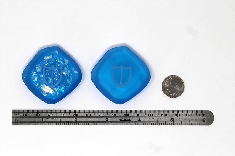 Puede incluir: Dos dados de resina de color azul, con forma de diamante, con un dise&ntilde;o de escudo blanco. Los dados miden aproximadamente 9 cm de di&aacute;metro. Se incluye una regla para la escala.