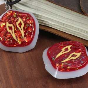 May include: Two red and white resin pendants with gold glitter and a gold design. The pendants are shaped like a rounded rectangle and have a white border. The pendants are on a wooden surface.