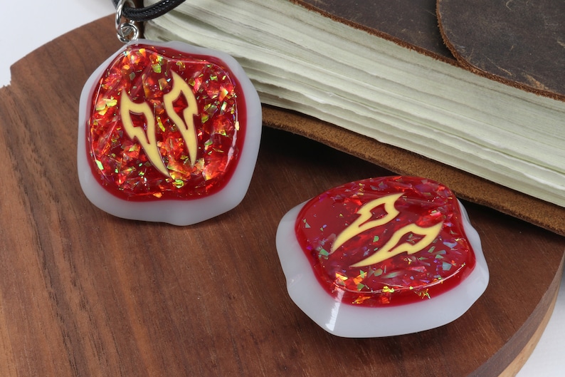 May include: Two red and white resin pendants with gold glitter and a gold design. The pendants are shaped like a rounded rectangle and have a white border. The pendants are on a wooden surface.