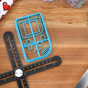 May include: A blue plastic cookie cutter shaped like a handheld video game console. The cookie cutter is on a wooden surface with a black ruler and a blue spatula.