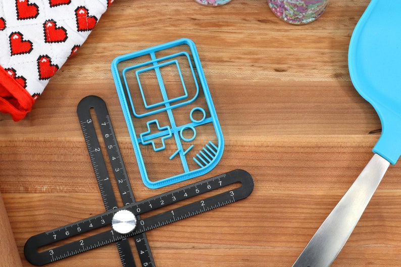 May include: A blue plastic cookie cutter shaped like a handheld video game console. The cookie cutter is on a wooden surface with a black ruler and a blue spatula.