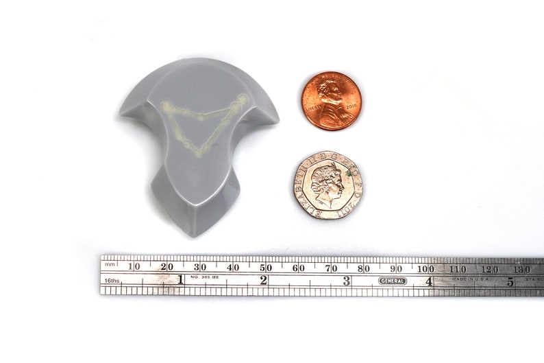 May include: A silver-colored, three-dimensional, abstract sculpture with a geometric shape. The sculpture is about 2 inches tall and 2 inches wide. It is sitting on a white surface next to a ruler and two coins.