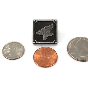 May include: A silver metal pin with a black background and a silver anvil design. The pin is about the size of a US quarter.