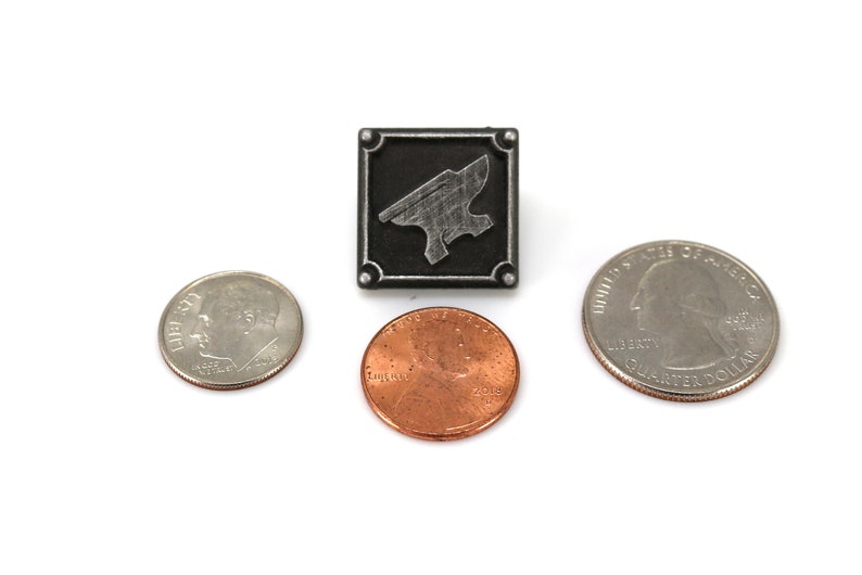 May include: A silver metal pin with a black background and a silver anvil design. The pin is about the size of a US quarter.
