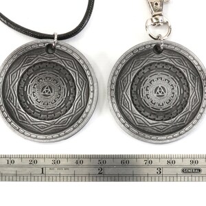 May include: Two silver-toned pendants, one on a black cord and the other on a silver chain, feature a detailed design of a stylized triangle within a circle. The pendants are approximately 2 inches in diameter.