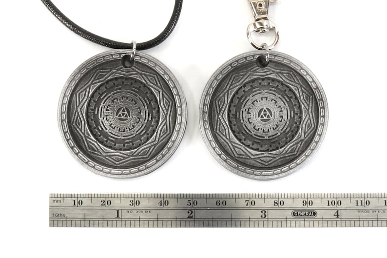 May include: Two silver-toned pendants, one on a black cord and the other on a silver chain, feature a detailed design of a stylized triangle within a circle. The pendants are approximately 2 inches in diameter.