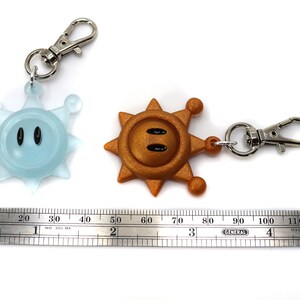 May include: Two keychains featuring a blue and a gold star-shaped character with a face. The characters have black eyes and a small mouth. The keychains are attached to silver metal rings and clips.