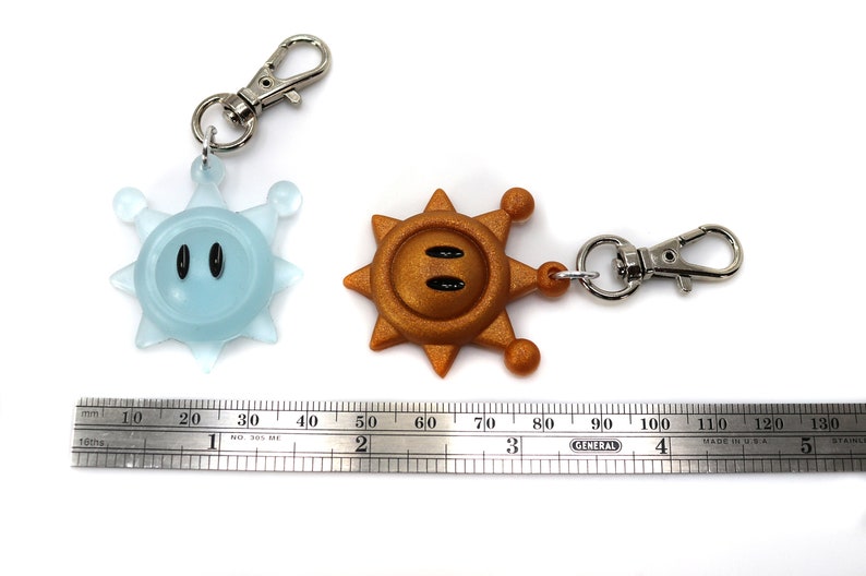 May include: Two keychains featuring a blue and a gold star-shaped character with a face. The characters have black eyes and a small mouth. The keychains are attached to silver metal rings and clips.
