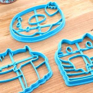 May include: Three blue plastic cookie cutters shaped like a doughnut, a cat, and a cat wearing a hat. The doughnut cutter has a hole in the centre and a strawberry on top. The cat cutter has a striped pattern. The cat wearing a hat cutter has a striped pattern and a hat on its head.