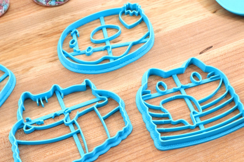 May include: Three blue plastic cookie cutters shaped like a doughnut, a cat, and a cat wearing a hat. The doughnut cutter has a hole in the centre and a strawberry on top. The cat cutter has a striped pattern. The cat wearing a hat cutter has a striped pattern and a hat on its head.