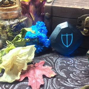May include: A blue, faceted, gemstone with a shield design etched into it. The gemstone is sitting on a dark, patterned surface with other decorative items.