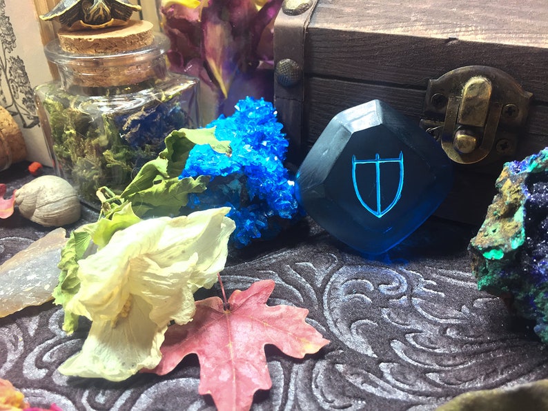May include: A blue, faceted, gemstone with a shield design etched into it. The gemstone is sitting on a dark, patterned surface with other decorative items.