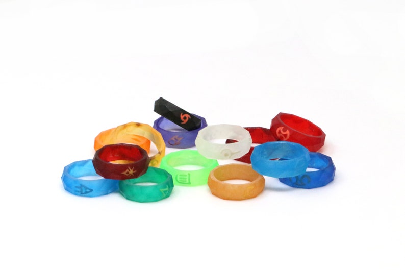 May include: A collection of 12 colorful silicone rings with different symbols. The rings are stacked and arranged in a semi-circle on a white background.