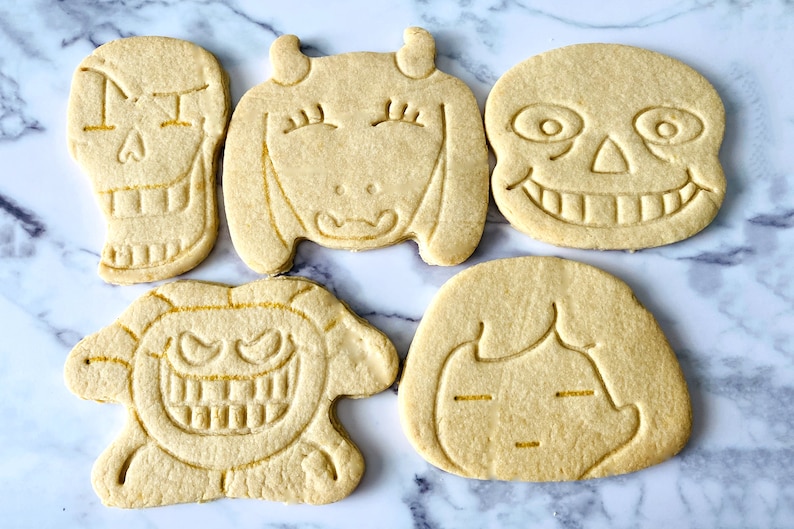 May include: Five decorated sugar cookies, each featuring a different character from the video game "Undertale". The cookies are light brown and have a slightly rough texture. The characters are Sans, Papyrus, Toriel, Asriel, and Frisk.