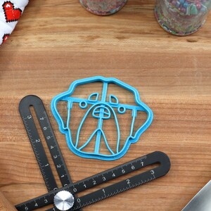 May include: A blue plastic cookie cutter shaped like a dog's face. The cutter is on a wooden surface with a ruler and a rolling pin.
